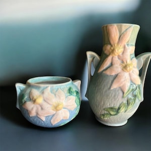 May include: Two ceramic vases in a floral design. The vase on the left is a small, round bowl with handles, and the vase on the right is a taller, handled vase. Both vases are light blue with pink and yellow flower details.