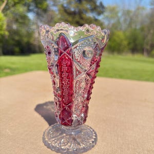 May include: A ruby red and clear glass vase with a detailed, textured pattern. The vase has a flared top and a round, decorative base. The glass catches the light, creating a sparkling effect. The vase is likely vintage or antique.