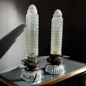 May include: Two vintage clear glass table lamps. Each lamp has a textured, cylindrical body and a ribbed, conical top. The lamps sit on a clear, faceted base and have brown electrical cords. The lamps are on a reflective surface.