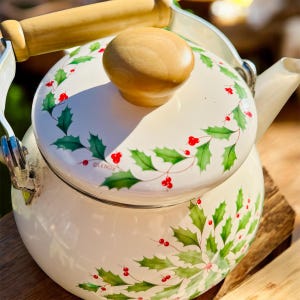 May include: Cream-colored enamel tea kettle with a wooden handle and lid knob. The kettle is decorated with green holly leaves and red berries. The handle and lid knob are made of light-colored wood. The kettle has a silver-colored spout and handle attachments.
