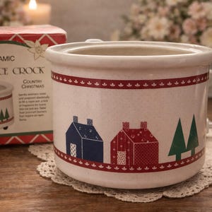 May include: A ceramic fragrance crock with a Country Christmas theme. The off-white crock features a red band with small floral accents and a repeating pattern of blue and red houses and green trees. The box next to it has the same design.