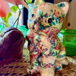 May include: A ceramic cat figurine with a floral pattern in pink, red, and green. The cat is sitting upright with its ears perked up. The figurine is on a woven surface.