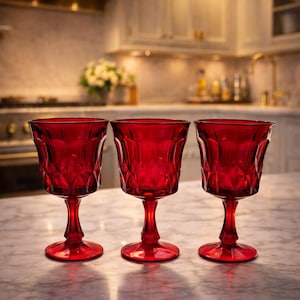 May include: Three ruby red wine glasses with a diamond pattern sit on a marble countertop. The stemware has a classic design, with a rounded bowl and a slender stem. The background shows a kitchen with white cabinets and a stainless steel range hood.
