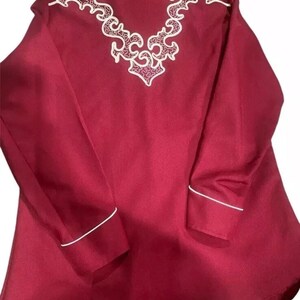 Puede incluir: Una camisa de manga larga de color burdeos con bordados blancos en la espalda. La camisa tiene ribetes blancos alrededor del cuello y los pu&ntilde;os.