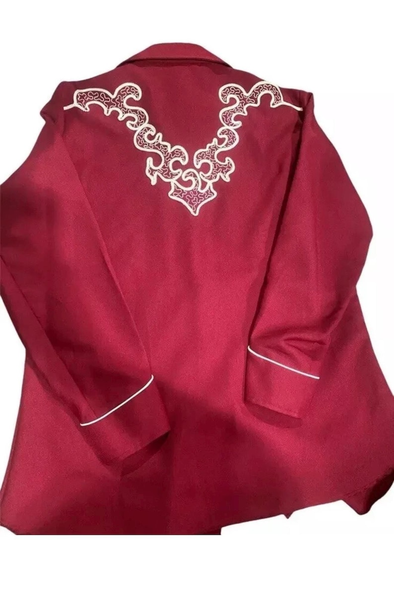 Puede incluir: Una camisa de manga larga de color burdeos con bordados blancos en la espalda. La camisa tiene ribetes blancos alrededor del cuello y los pu&ntilde;os.