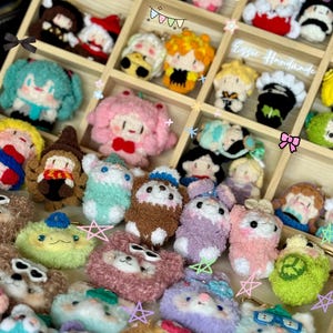 May include: A collection of handmade crochet keychains in various colors and character designs. The keychains are displayed in a wooden organizer. The text "Essie Handmade" is visible on the top right.