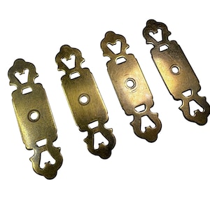 Op de afbeelding: Vier antieke decoratieve hardware-onderdelen in messingkleur. Elk stuk heeft een rechthoekig midden met een cirkelvormig gat en sierlijke, symmetrische uitgesneden ontwerpen aan de uiteinden. De hardware is waarschijnlijk voor meubels of kasten.