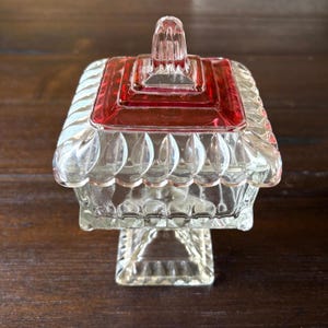 May include: A clear glass candy dish with a red lid. The dish has a square base and a decorative, scalloped edge. The lid has a square handle. The dish is likely vintage and would be a nice addition to a home decor collection.