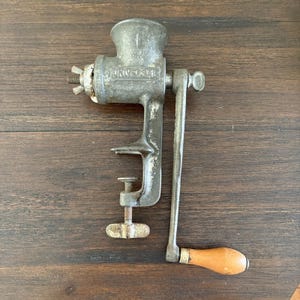May include: Vintage metal Universal meat grinder with a wooden handle. The grinder is silver with visible wear and tear, featuring a clamp for secure attachment. The handle is made of wood and is light brown. The word "UNIVERSAL" is embossed on the side.
