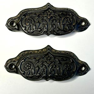 May include: Two black metal drawer pulls featuring an ornate, raised floral and scrollwork pattern. Each handle has a central pointed arch and two circular screw holes on the outer ends, with a textured background within the decorative design.