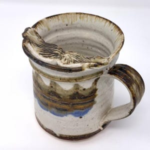 May include: Handmade ceramic mug with a unique, textured rim detail. The mug has a cream-coloured base with brown, blue, and tan accents. The handle is brown and the rim has a decorative, carved design. A rustic, handcrafted piece.