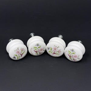 May include: Set of four white ceramic cabinet knobs. Each knob features a floral design with pink flowers, green leaves, and the text "Rosa Canina". The knobs have silver metal stems.