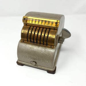 Op de afbeelding: Vintage grijze en messing mechanische rekenmachine met een hendel en meerdere nummerwielen. De rekenmachine heeft een rechthoekige vorm met een helder plastic venster en is gemarkeerd met "Presto". Hij staat op een donkerbruine basis.