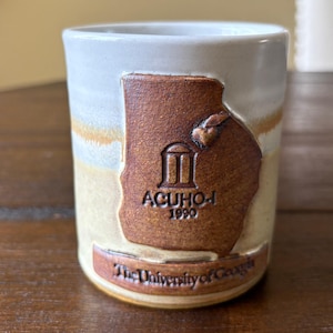 May include: A ceramic mug with a light beige and brown glaze. The mug features a raised brown design of the state of Georgia with the text "ACUHO-I 1990" and "The University of Georgia".
