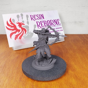 May include: A gray 3D printed miniature of a hooded figure holding a bow and arrow, standing on a round base. The miniature is on a wooden surface. The text "RESIN REBORNE" is visible on a white card behind the miniature.