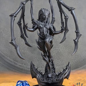 May include: A black and gray 3D printed miniature of a female spider creature with long, spindly legs and a menacing expression. The creature is standing on a base with a jagged, claw-like design.
