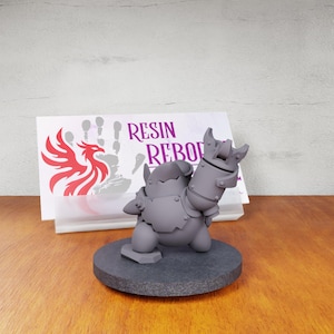 May include: A gray, unpainted resin miniature of a stout, armored creature holding a large cannon-like weapon, standing on a circular base. Behind it is a white rectangular card featuring a red phoenix logo and the text RESIN REBORN.