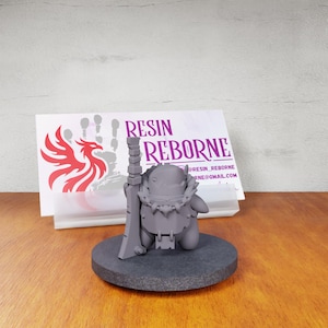 May include: A gray resin miniature figure of a creature wearing a spiked collar and holding a large rectangular sword, standing on a circular base. Behind the figure is a white card with the text RESIN REBORNE and a red phoenix logo.
