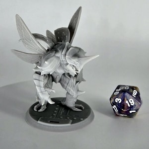 May include: A gray, insectoid fantasy miniature figure with large wings, horns, and a detailed exoskeleton, standing on a circular base. A multi-sided die with purple and white markings is next to the figure, suggesting a tabletop game.