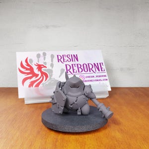 May include: A gray, unpainted resin figurine of a stout, armored character holding a shield and a mace, mounted on a circular base. Behind it sits a white business card with the text RESIN REBORNE, a red phoenix logo, and contact details.