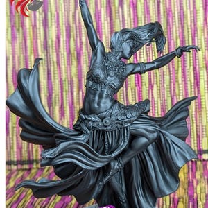 May include: A black painted miniature figurine of a woman in a flowing dress, dancing with her arms outstretched. The figurine is posed on a circular base with a purple and white dice.