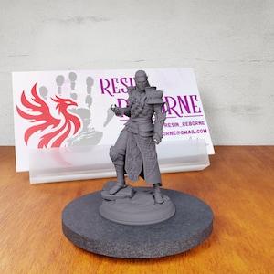 May include: A gray 3D printed miniature of a fantasy character wearing a detailed outfit and holding a staff. The miniature is standing on a round base. The background includes a business card with the text "Resin_Reborne" and a website address.