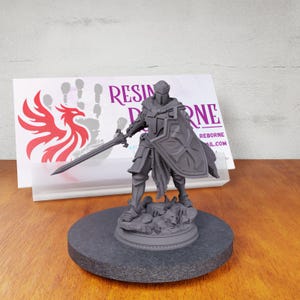 May include: Gray painted miniature figurine of a knight holding a sword and shield. The knight is standing on a rocky base. The figurine is on a black circular base. The background includes a business card with the text "RESIN REBORNE" and a red phoenix logo.