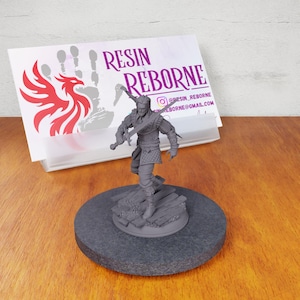 May include: A gray 3D printed miniature of a warrior figure holding a sword and shield. The figure is standing on a wooden base. The miniature is on a gray circular base. The background includes a business card with the text "RESIN REBORNE" and a social media handle.