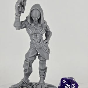 May include: A gray plastic miniature figurine of a female character wearing a hooded outfit and holding a gun. The figurine is standing on a round base with a textured surface. A purple 20-sided die is in the foreground.