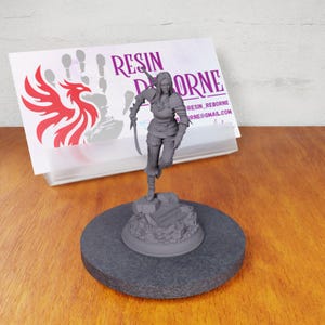 May include: A gray, unpainted miniature figurine of a female warrior holding a sword and standing on a rocky base. The figurine is on a black circular base.