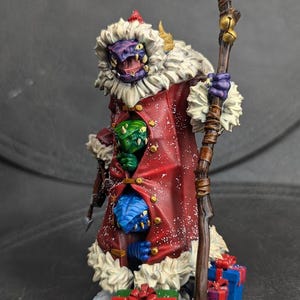 May include: A purple and white painted miniature figure of a dragon wearing a red coat with white fur trim and a Santa hat. The dragon is holding a wooden staff with bells. There are three smaller dragons peeking out from under the coat. The figure is standing on a base with snow and wrapped presents.