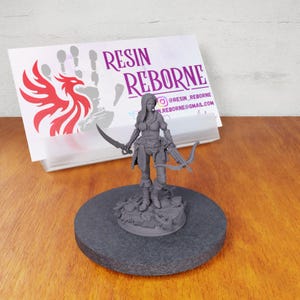 May include: Gray 3D printed miniature of a female figure holding a bow and arrow and a sword. The figure is standing on a round base with a detailed design. The miniature is next to a business card with the text "Resin Reborne" and social media information.