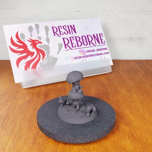 May include: A gray 3D printed miniature of a mushroom person standing on a stone base with smaller mushrooms around it. The miniature is on a gray circular base.