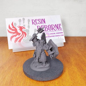 May include: A gray, 3D printed miniature of a dwarf warrior holding a large axe and a shield. The miniature is standing on a round base. The miniature is a fantasy character.