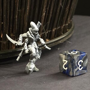 May include: A gray miniature figure of a futuristic warrior with two curved blades and a detailed armored design. Beside it is a six-sided die with white numbers on a blue and gray marbled surface. The scene is set on a dark surface.
