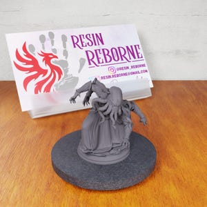 May include: A gray, 3D printed miniature of a humanoid figure with tentacles, wearing a robe. The figure is posed in a dynamic stance, with one hand raised and the other extended. The miniature is sitting on a black circular base. The business card in the background reads "Resin Reborn" with a website and Instagram handle.