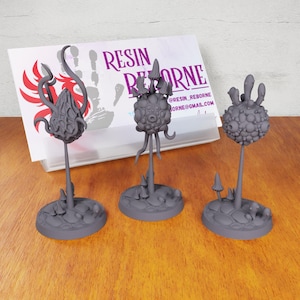 May include: Three gray resin miniature fantasy plant sculptures on round bases. Each plant has a unique design with a bulbous top and multiple stems. The bases are textured to resemble dirt and rocks. The sculptures are labeled "Resin Reborne" on a card in the background.