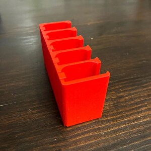 Puede incluir: Un organizador de pl&aacute;stico rojo con cinco ranuras para guardar objetos peque&ntilde;os. El organizador est&aacute; hecho de pl&aacute;stico rojo brillante y tiene un acabado liso y brillante.