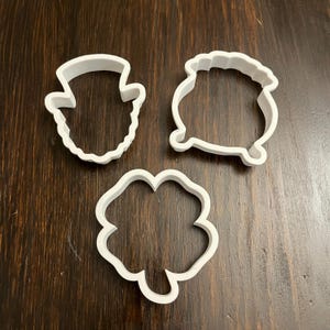 May include: Three white plastic cookie cutters: a leprechaun hat, a pot of gold, and a four-leaf clover.