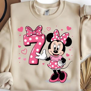 May include: A beige sweatshirt featuring a pink Minnie Mouse graphic. Minnie Mouse is wearing a pink polka dot dress and bow. A large pink number 7 with white polka dots is next to Minnie, with pink hearts scattered around.