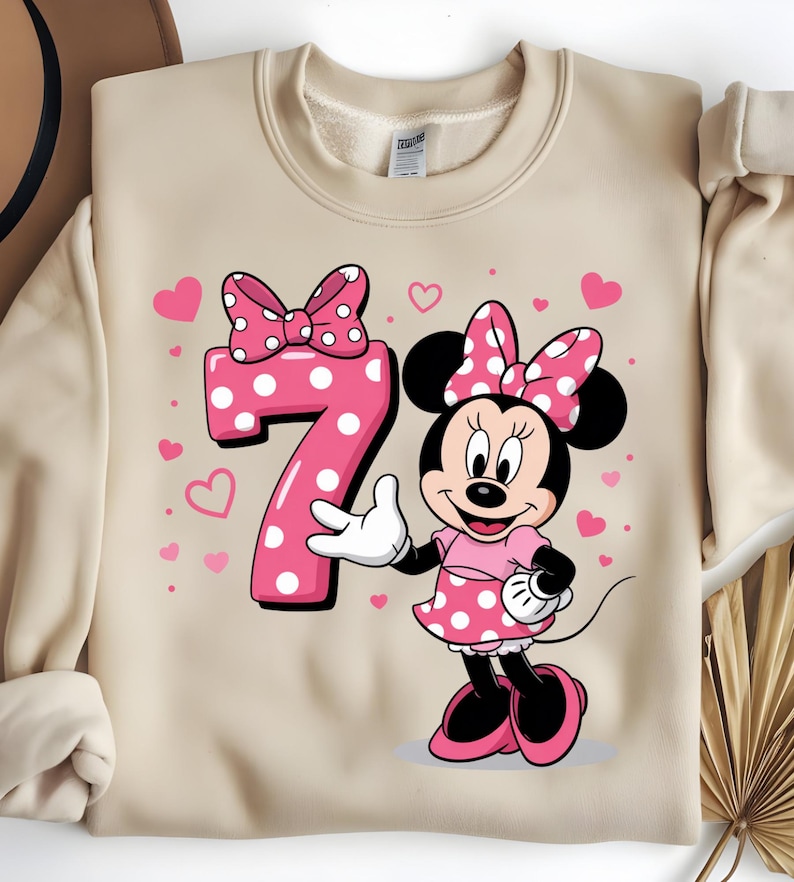 May include: A beige sweatshirt featuring a pink Minnie Mouse graphic. Minnie Mouse is wearing a pink polka dot dress and bow. A large pink number 7 with white polka dots is next to Minnie, with pink hearts scattered around.