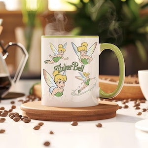 Pode incluir: Caneca de cerâmica branca com ilustrações da Sininho, uma alça e borda verdes. A caneca está sobre um descanso de madeira, rodeada de grãos de café. A caneca tem o texto "Tinker Bell".