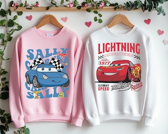 May include: Two pink and white sweatshirts with a Cars theme. The pink sweatshirt features a blue car with the text "Sally" and the white sweatshirt features a red car with the text "Lightning McQueen".