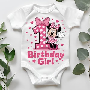 May include: White baby bodysuit with a pink number 1, Minnie Mouse graphic, and the text "Birthday Girl." The number 1 has a polka dot design and is next to a bow. Pink hearts are scattered around the design.