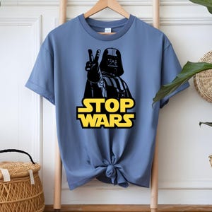 May include: A blue t-shirt featuring a black graphic of Darth Vader making a peace sign. The text "STOP WARS" is in yellow block letters below the image. The shirt is hanging on a wooden hanger.