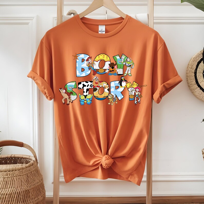 Pu&ograve; includere: T-shirt arancione con la scritta "BOY STORY" in lettere colorate in stile cartoon. Le lettere sono riempite con immagini di personaggi. La maglietta ha le maniche arrotolate e un nodo sul davanti.