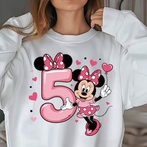 May include: White sweatshirt featuring a pink number 5 with a Minnie Mouse graphic. Minnie Mouse is wearing a pink polka dot dress and holding a heart-shaped balloon. Pink hearts and black dots are scattered around the design.