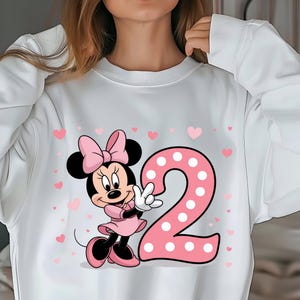 May include: White sweatshirt featuring a Minnie Mouse graphic with a pink bow, dress, and shoes. A large pink number 2 with white polka dots is next to Minnie. Small pink hearts are scattered around the design.