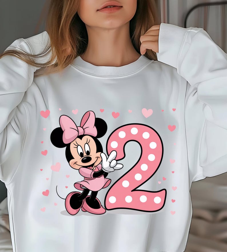 May include: White sweatshirt featuring a Minnie Mouse graphic with a pink bow, dress, and shoes. A large pink number 2 with white polka dots is next to Minnie. Small pink hearts are scattered around the design.