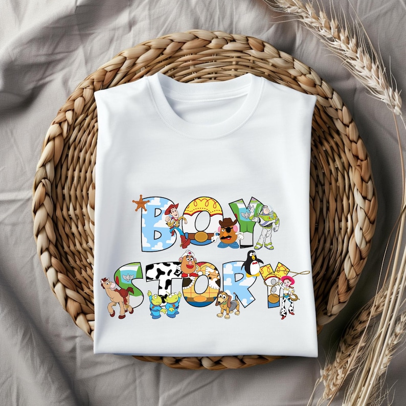 Pu&ograve; includere: T-shirt bianca con la scritta "BOY STORY" in lettere colorate e giocose. Il design incorpora personaggi dei cartoni animati. La maglietta &egrave; esposta su un cestino intrecciato, con spighe di grano sullo sfondo.